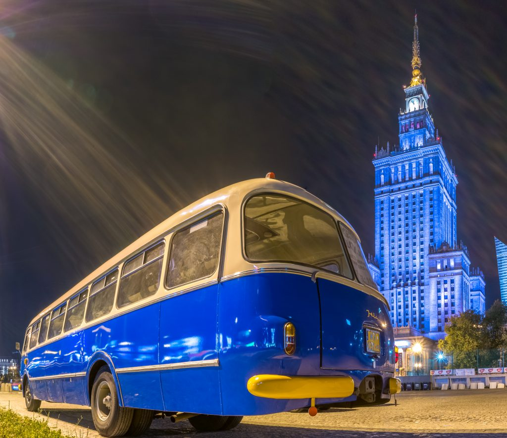 zdjęcie przedstawia biało-niebieski autobus ogórek na tle podświetlonego na niebiesko Pałacu Kultury i Nauki